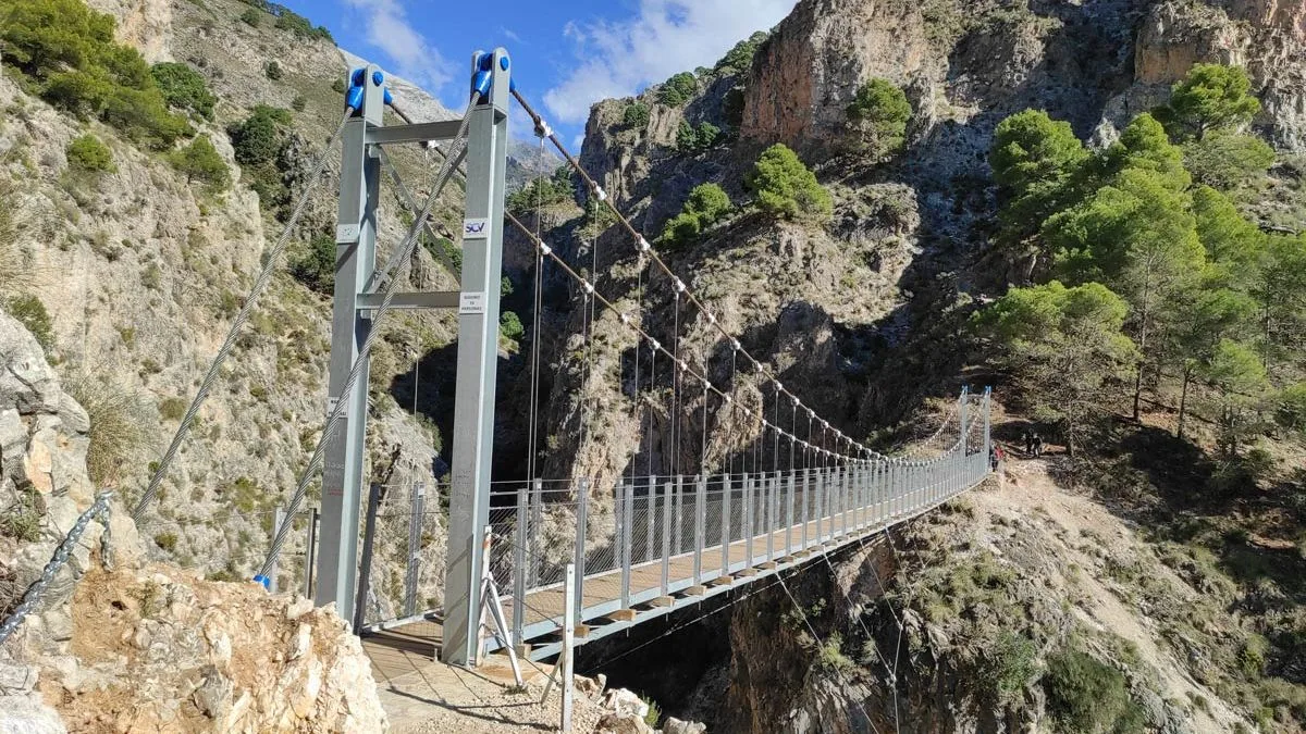 The El Saltillo suspension bridge seen from its near abutment, the steel walkway slung tower to tower across the gorge above the Río Almanchares with walkers waiting on the far side